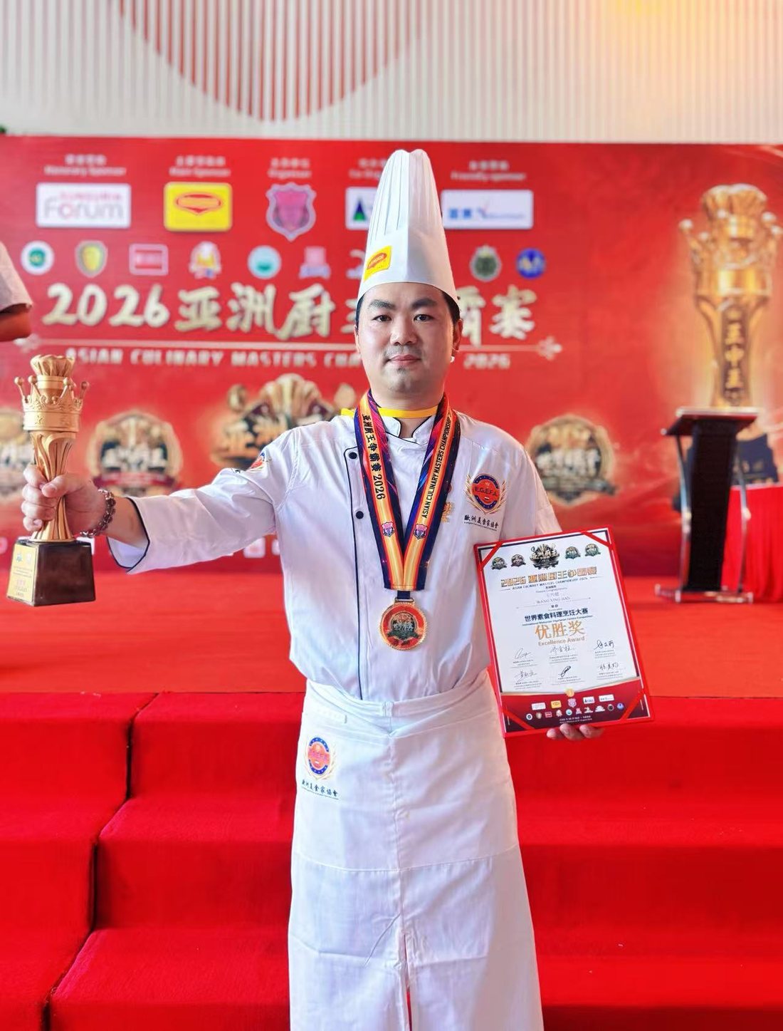 Chef Wang Xing Jian met zijn gouden beker en oorkonde voor de backdrop van de Asian Culinary Masters Championship 2026